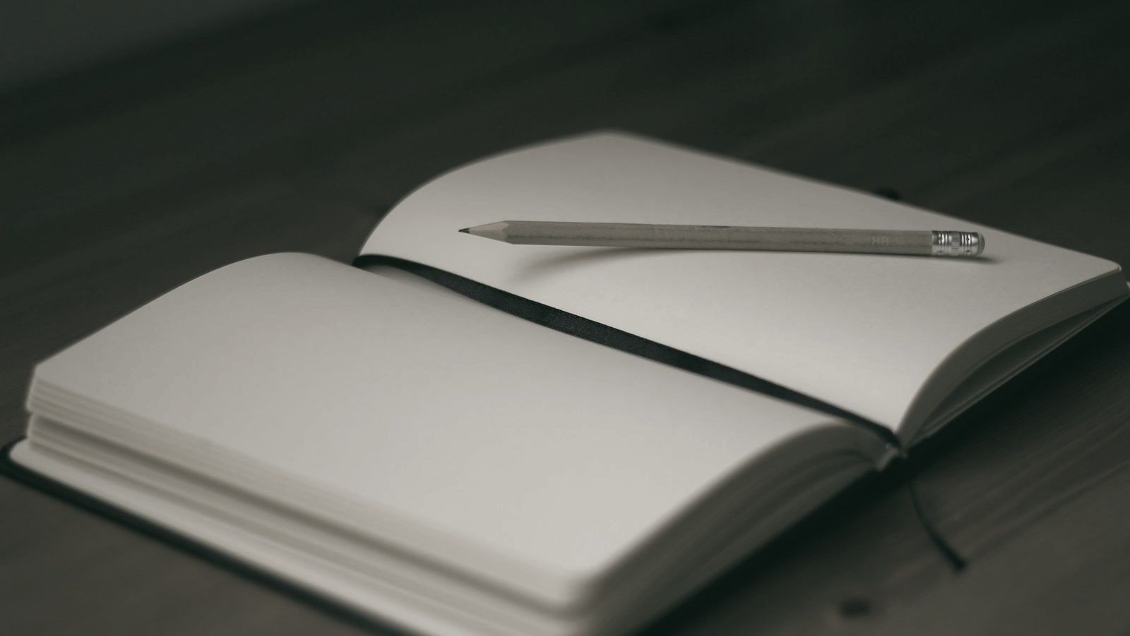 Cuaderno abierto con las páginas en blanco y un lápiz apoyado encima, sobre una mesa de madera.