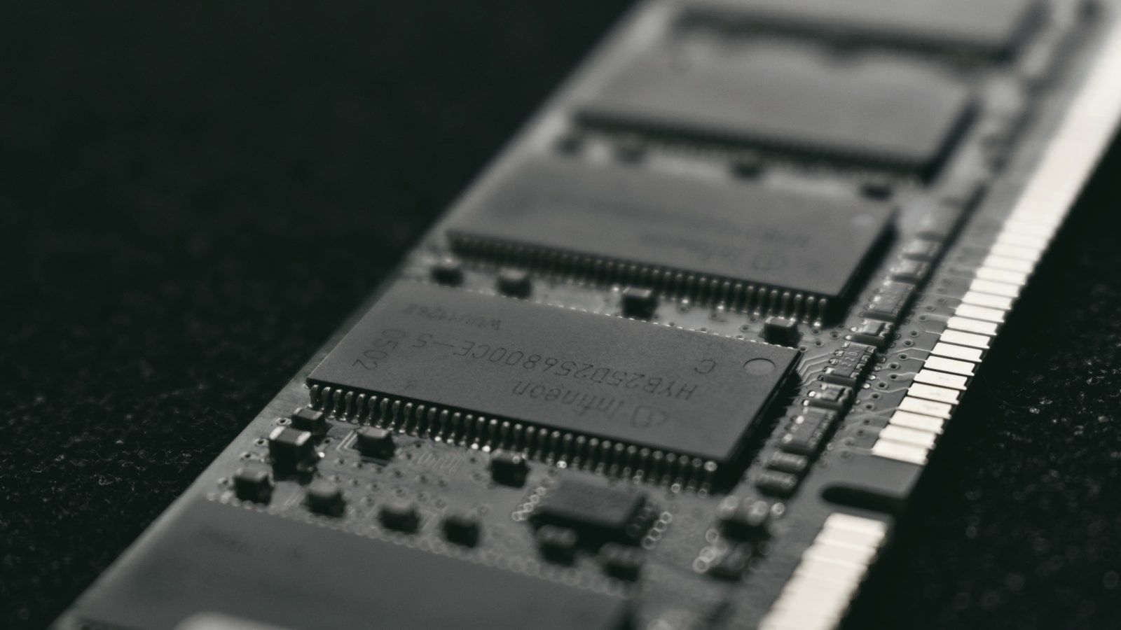 Close-up of a RAM module on a black background, with the chips and gold edge contacts in focus along a diagonal.