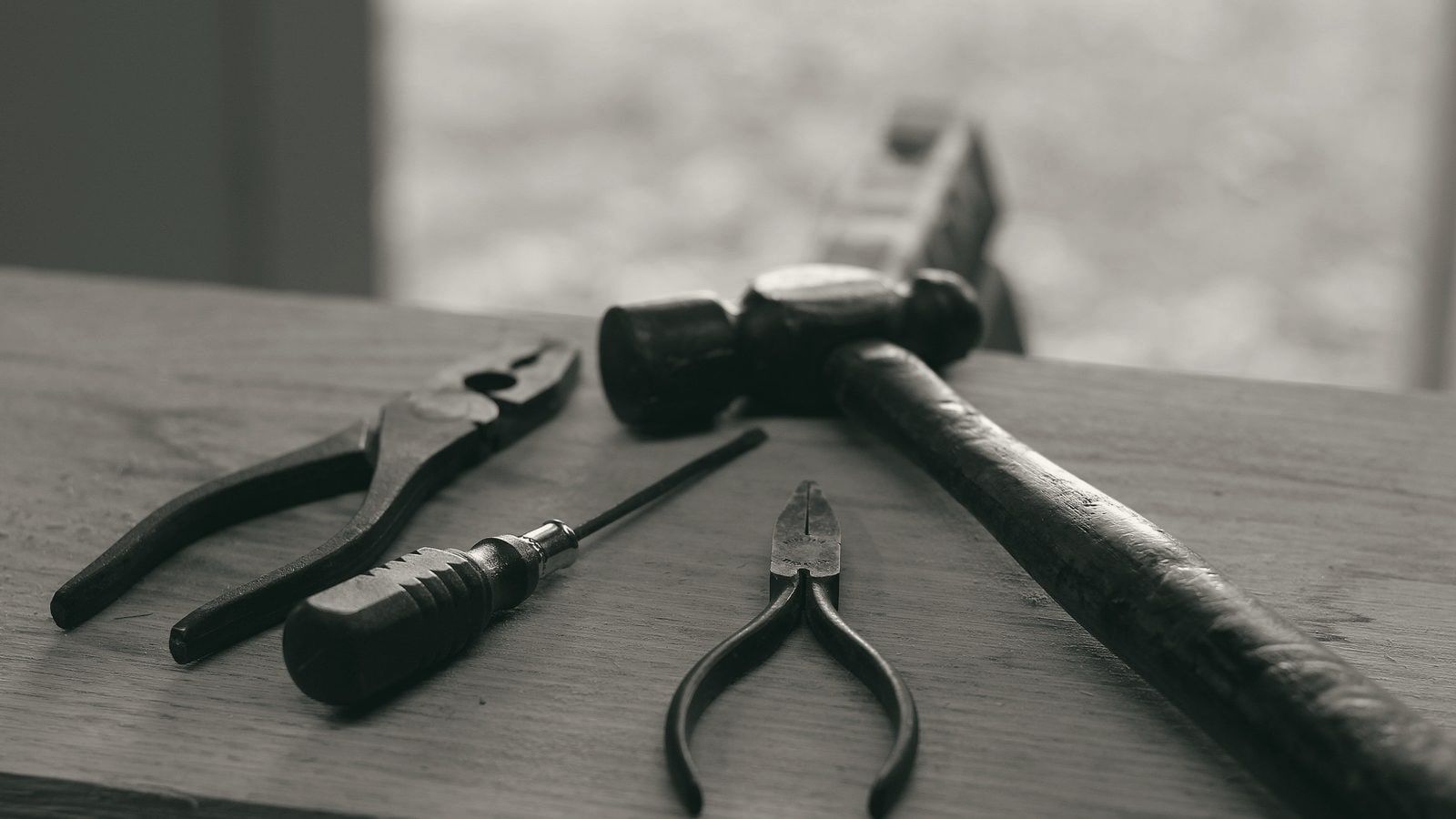 Four hand tools on a wooden board: pliers, a screwdriver, needle-nose pliers, and a hammer.